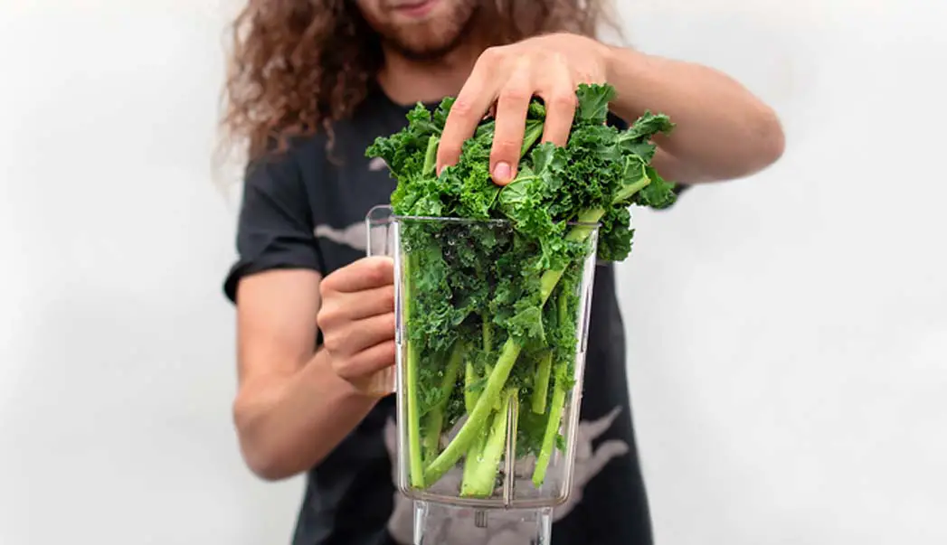 A man packing a blender with rich green kales leaves and stems.  