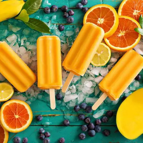 Colorful display of delicious vegan popsicles for a homemade popsicles recipe with fresh sliced oranges, blueberries, bananas, mango, and ice cubes on a bright aqua board.