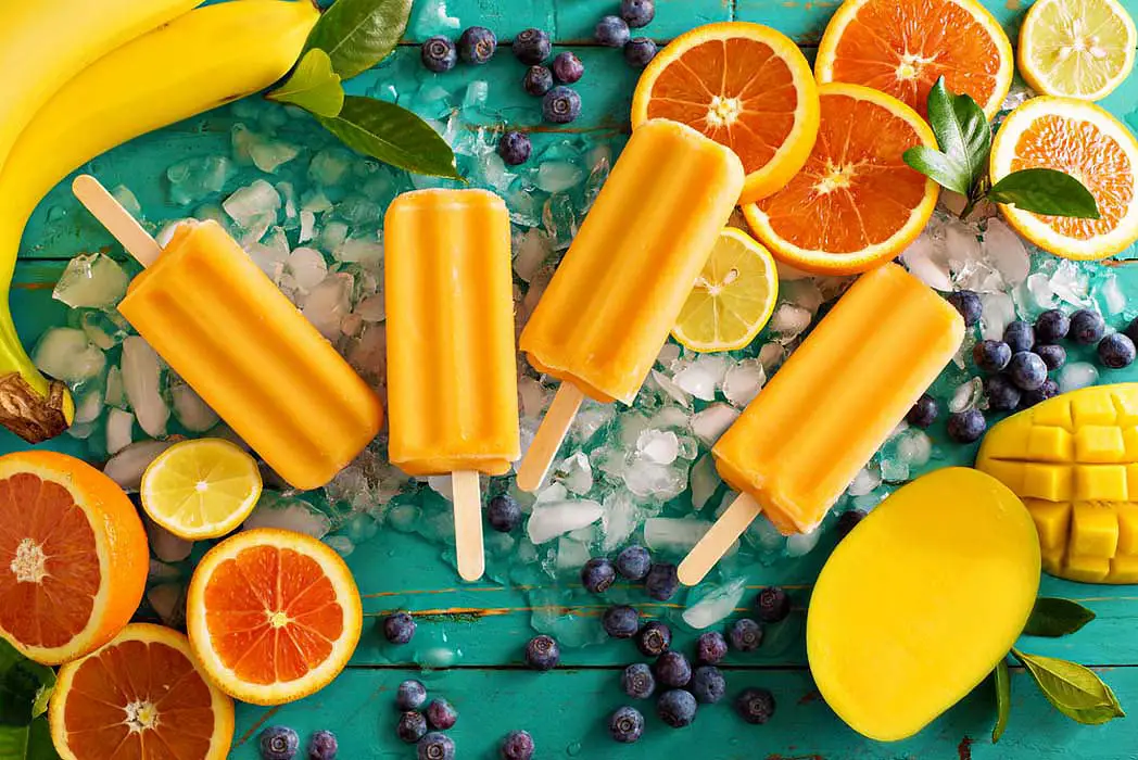 Colorful display of delicious vegan popsicles for a homemade popsicles recipe with fresh sliced oranges, blueberries, bananas, mango, and ice cubes on a bright aqua board.