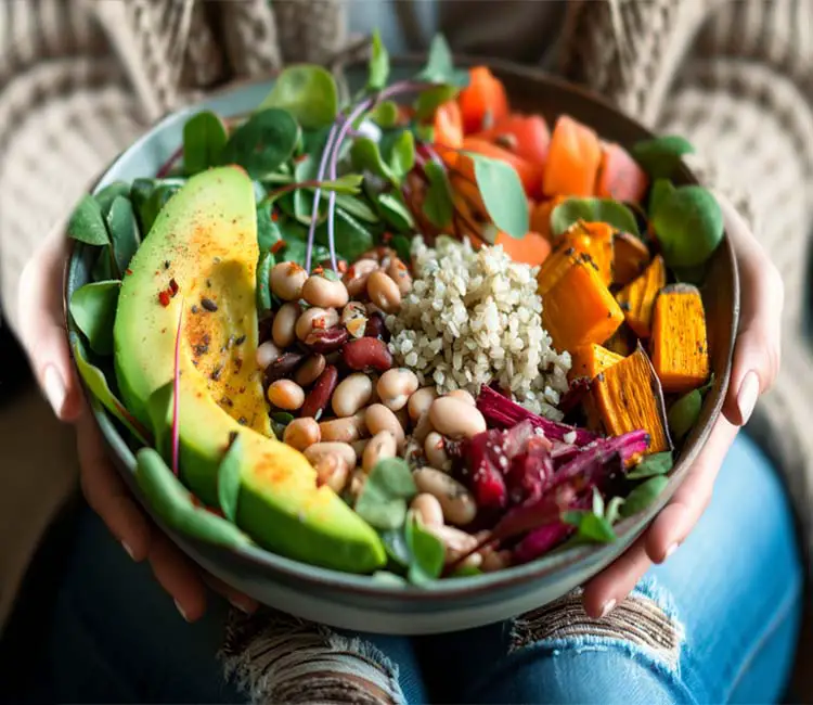 A woman in jeans and a fluffy sweater holding a stacked buddha bowl with avocados, beans, sprouts, veggies, sweet potatoes, and more.