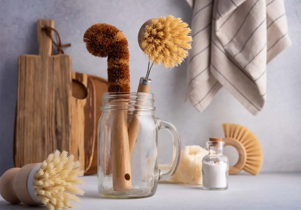 A large glass jar and kitchen counter full of eco-friendly cleaners, scrub brushes, and wooden cutting boards.