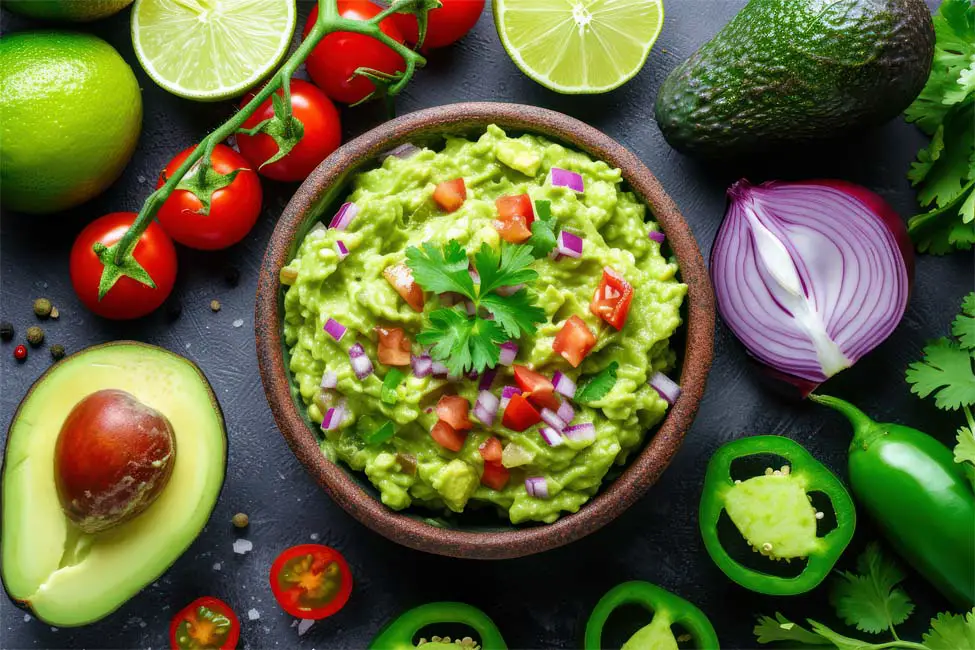 Top view of a rich bowl of organic guacamole topped with tomatoes, red onions, cilantro, and green peppers by a whole avocado and cherry tomatoes, peppers, onion, and fresh lime.