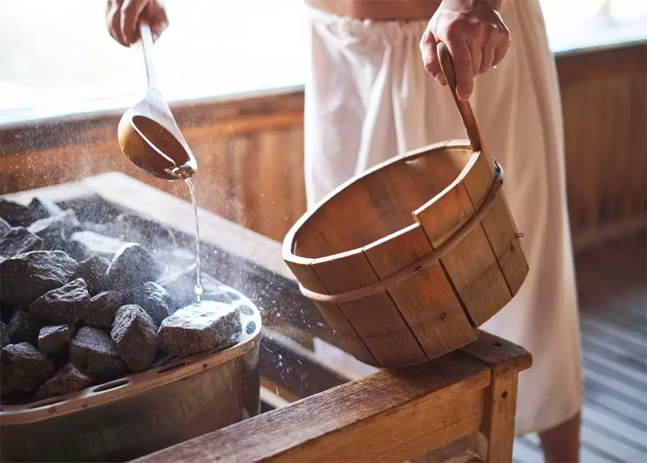 A man in a rustic wooden sauna ladling water on the stones from a cedar bucket.    