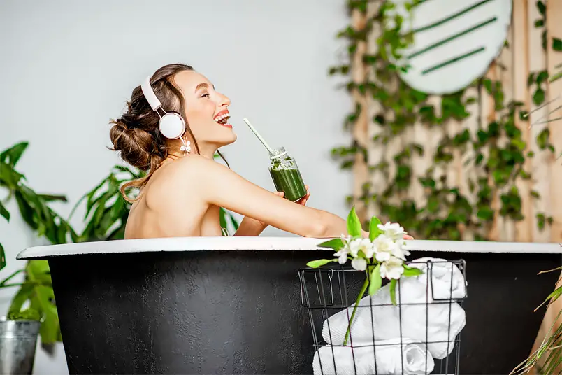 Happy smiling woman listening to music in the bathtub while detoxing on raw organic green juice.