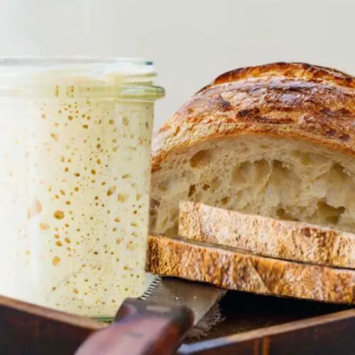 Buckwheat sourdough starter in a glass by a crusty loaf of sliced homemade sourdough bread.